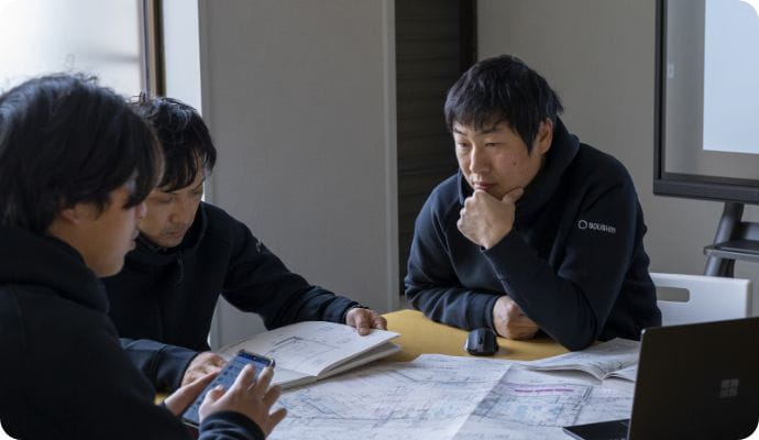 3人の男性が机上で資料とスマホを見ながら真剣に話し合っている。