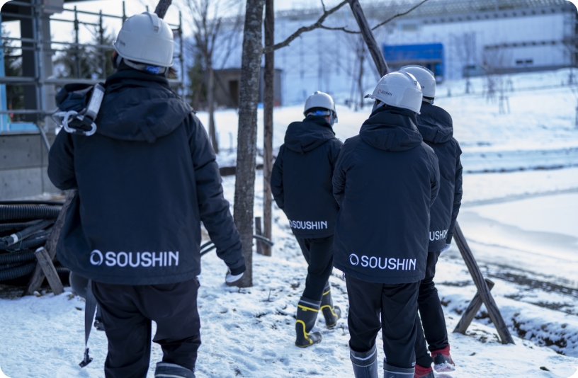 SOUSHINジャケットを着たヘルメット姿の作業員が雪道を歩く。