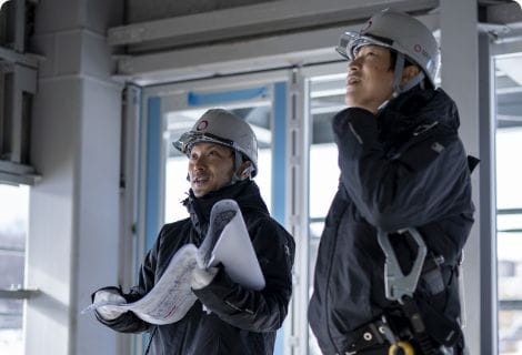 ヘルメットを被り図面を持つ二人の男性が建設現場を視察。